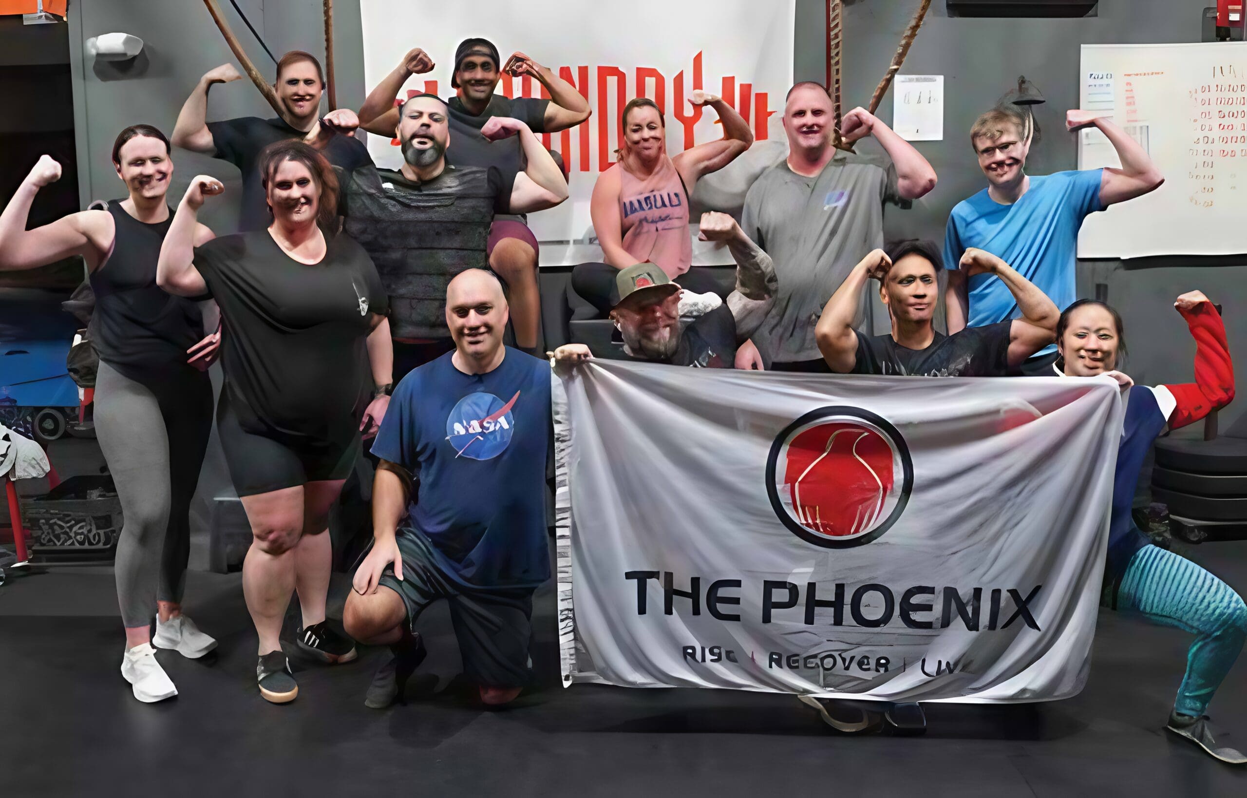 A diverse community at a gym poses together, flexing their arms and smiling. Two people kneel in front holding a banner that reads The Phoenix: Rise Recover Live, celebrating sober active living. Gym equipment is visible in the background.