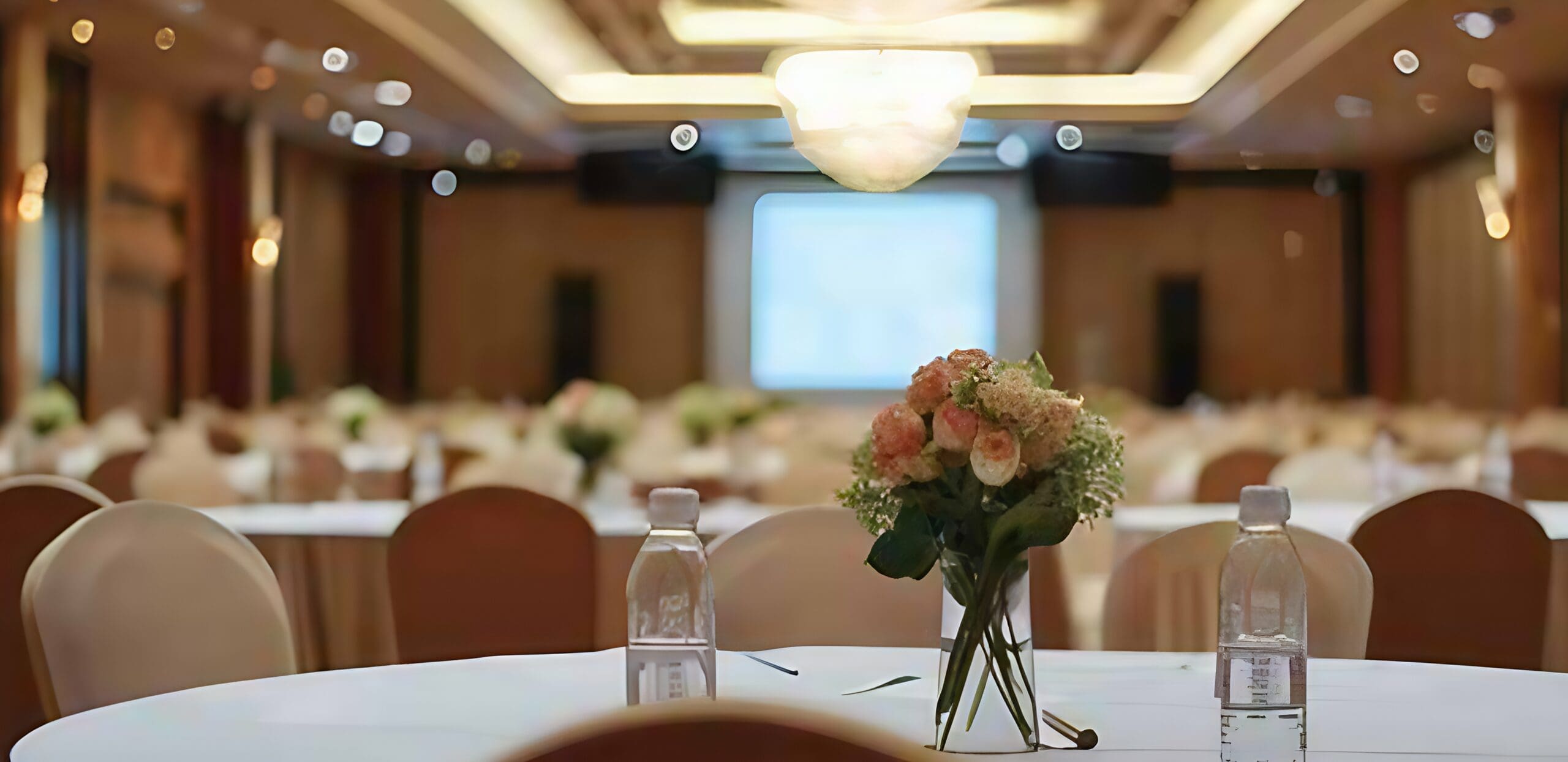 A banquet hall designed for inclusive gatherings, featuring round tables with white cloths, floral centerpieces, and water bottles. Soft lighting fills the room, with a large screen up front and chairs arranged for a welcoming atmosphere.