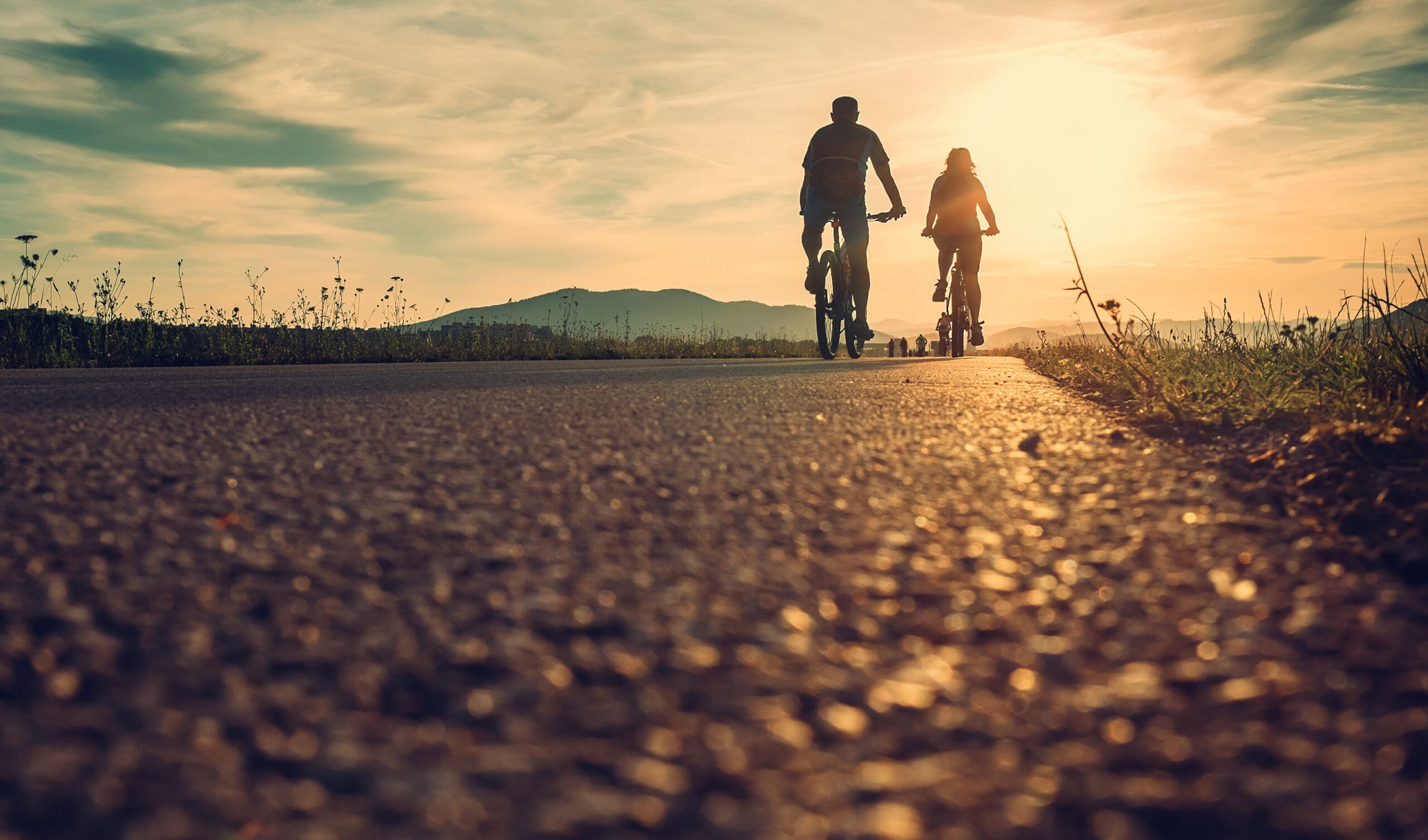 Two people ride bicycles on a paved path at sunset, their silhouettes visible against the bright sky and distant mountains—a peaceful moment that highlights recovery and supports physical well-being.