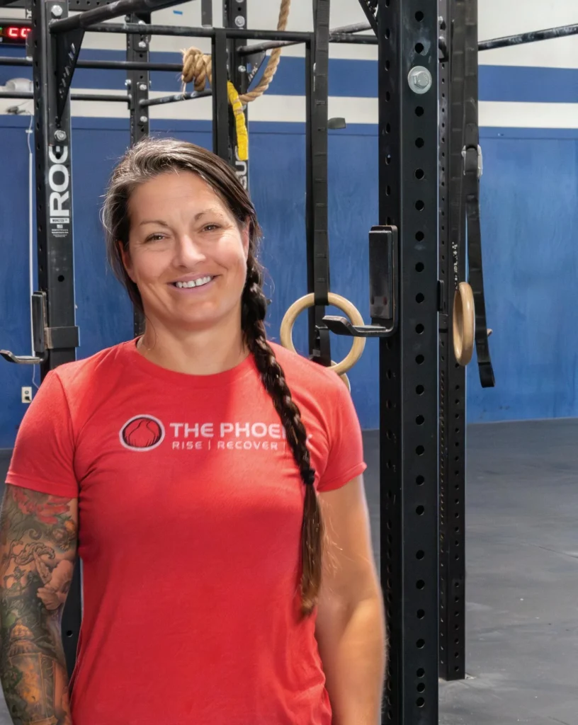 A woman with a long braid and a tattooed arm smiles while standing in a gym, wearing a red The Phoenix: Rise | Recover t-shirt. Gym equipment and blue walls are visible in the background.